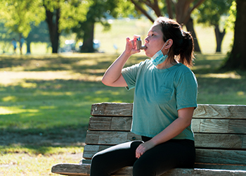 A woman sits on a bench in a park using an inhaler