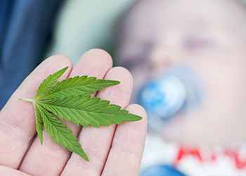A hand holds a marijuana leaf with a baby in the background