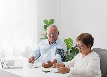 An older women sits at home and helps take her partner's blood pressure during a remote call to a health specialist