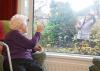 An older woman sits in her living room and waves to a carer outside
